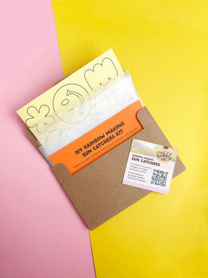 A DIY rainbow making sun catcher kit by The Bubbly Type, shown in a brown kraft envelope with printed templates and an orange instruction sheet, set against a split pink and yellow background.
