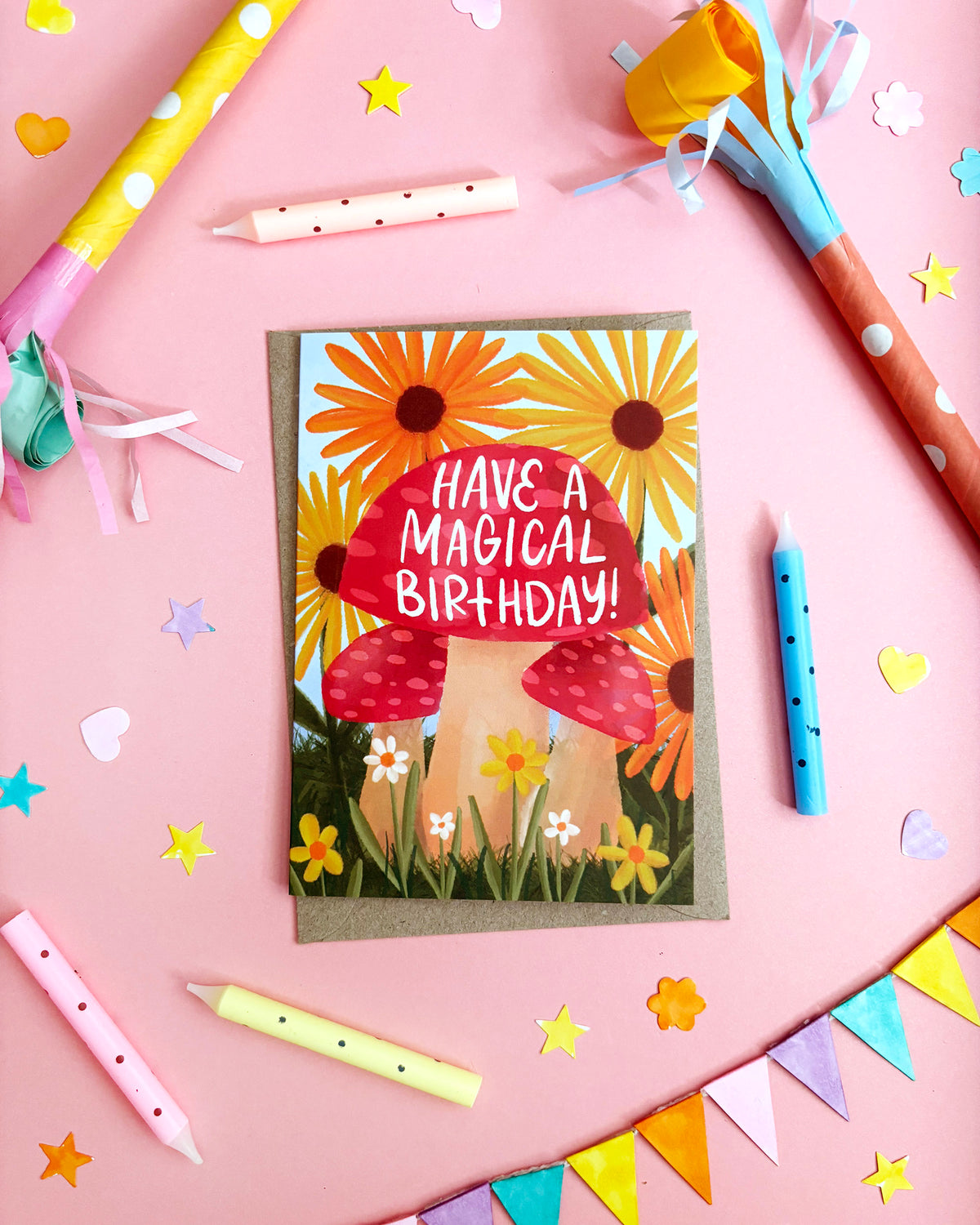 A  birthday card featuring two red mushrooms among yellow and orange flowers with the text 'HAVE A MAGICAL BIRTHDAY!' in white. The card is photographed from above on a pink background surrounded by party props including candles, paper horns, confetti, and bunting.