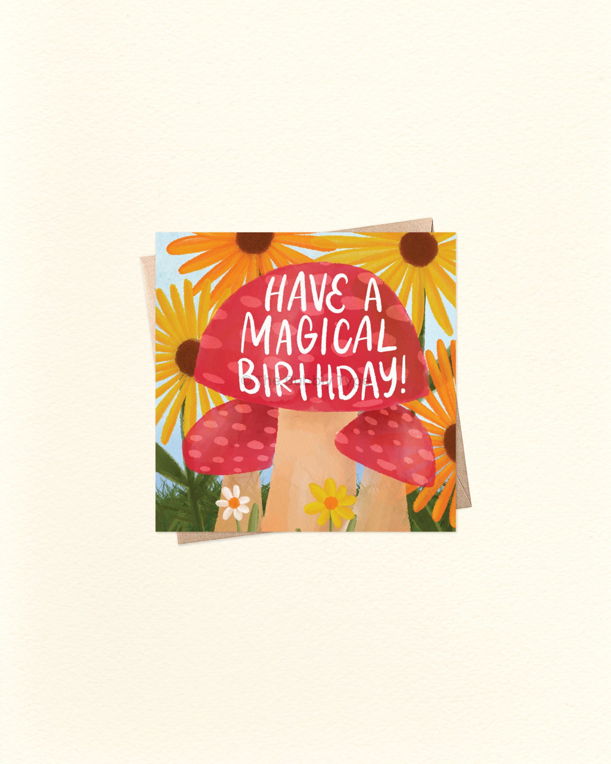 A square greeting card with a whimsical illustration of red-and-white mushrooms, yellow sunflowers, and small white flowers, with the text 'HAVE A MAGICAL BIRTHDAY!' in white lettering on the mushroom cap. The card is photographed on a warm off-white background.