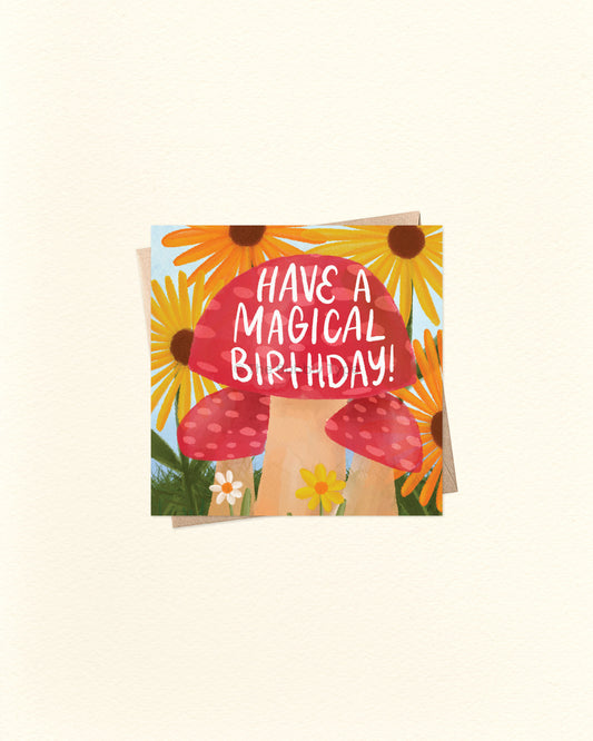 A square greeting card with a whimsical illustration of red-and-white mushrooms, yellow sunflowers, and small white flowers, with the text 'HAVE A MAGICAL BIRTHDAY!' in white lettering on the mushroom cap. The card is photographed on a warm off-white background.