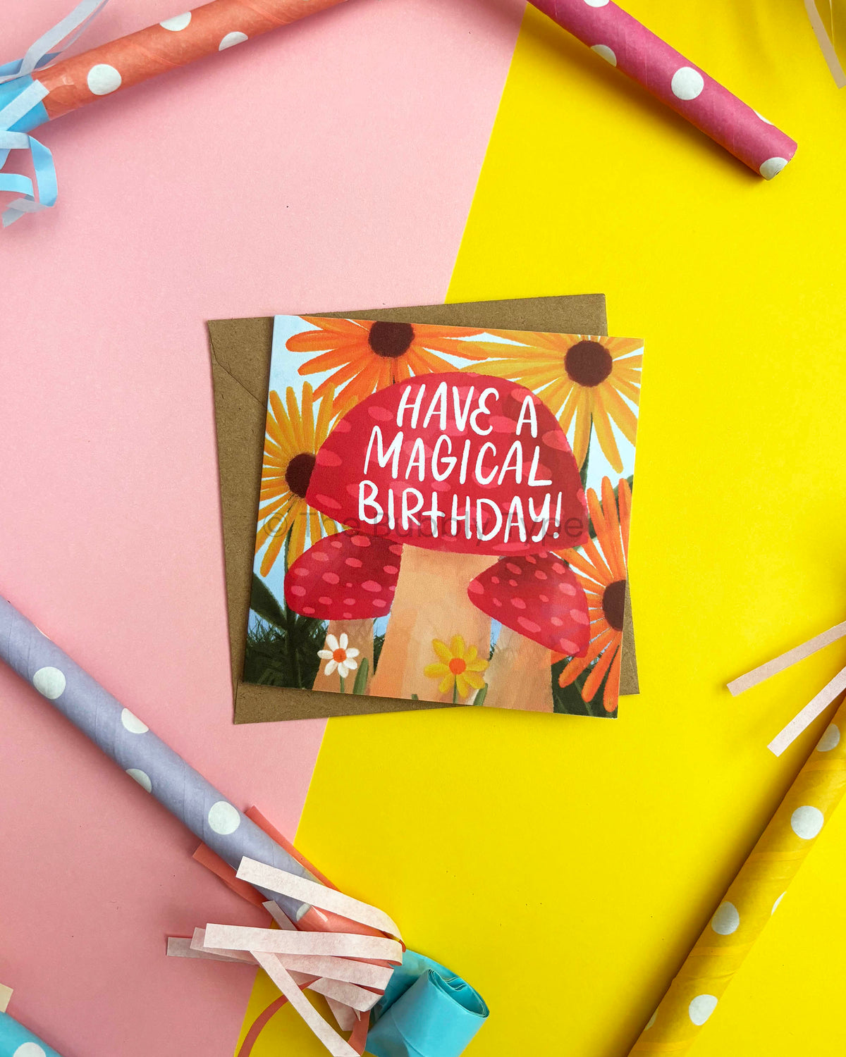 A square birthday card with a red mushrooms and yellow sunflower illustration and the text 'HAVE A MAGICAL BIRTHDAY!'. It lies on a brown envelope against a pink and yellow background with party horns and streamers around it.