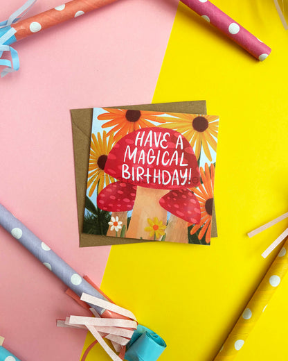 A square birthday card with a red mushrooms and yellow sunflower illustration and the text 'HAVE A MAGICAL BIRTHDAY!'. It lies on a brown envelope against a pink and yellow background with party horns and streamers around it.