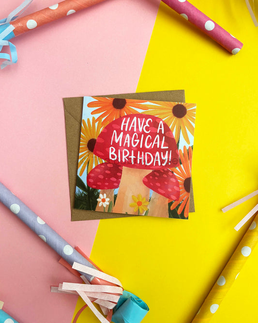 A square birthday card with a red mushrooms and yellow sunflower illustration and the text 'HAVE A MAGICAL BIRTHDAY!'. It lies on a brown envelope against a pink and yellow background with party horns and streamers around it.