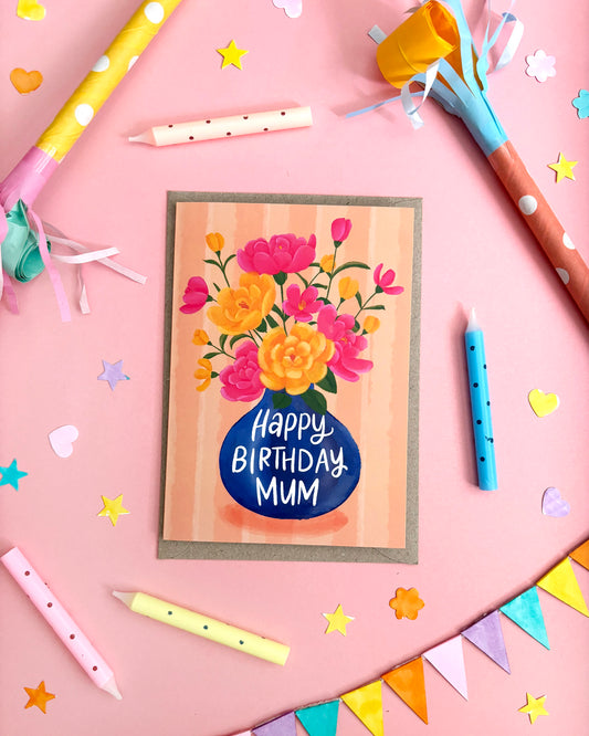 A birthday card with a floral bouquet and the text 'Happy Birthday Mum' in a blue circle, styled on a pink background with party props like candles, streamers, star cutouts, and bunting.