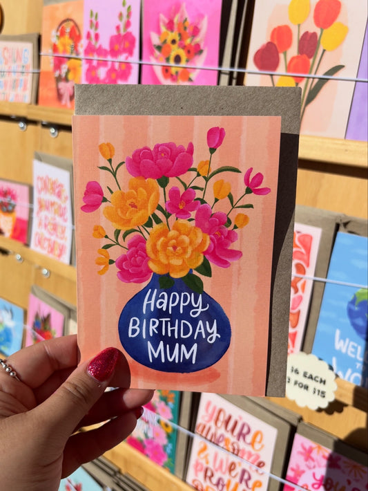 Hand holding a birthday card with flowers and 'Happy Birthday Mum' text, surrounded by other cards.
