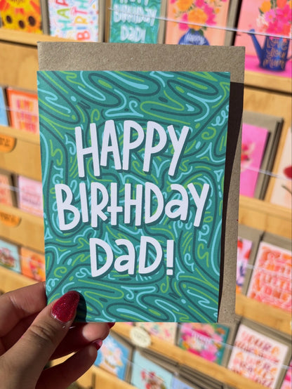 Hand holding a birthday card with 'Happy Birthday Dad!' text against a store background.
