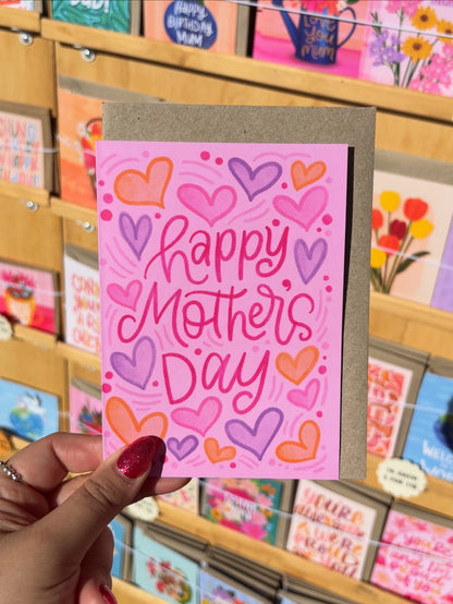 Hand holding a 'Happy Mother's Day' card with colourful hearts in front of a display of greeting cards.