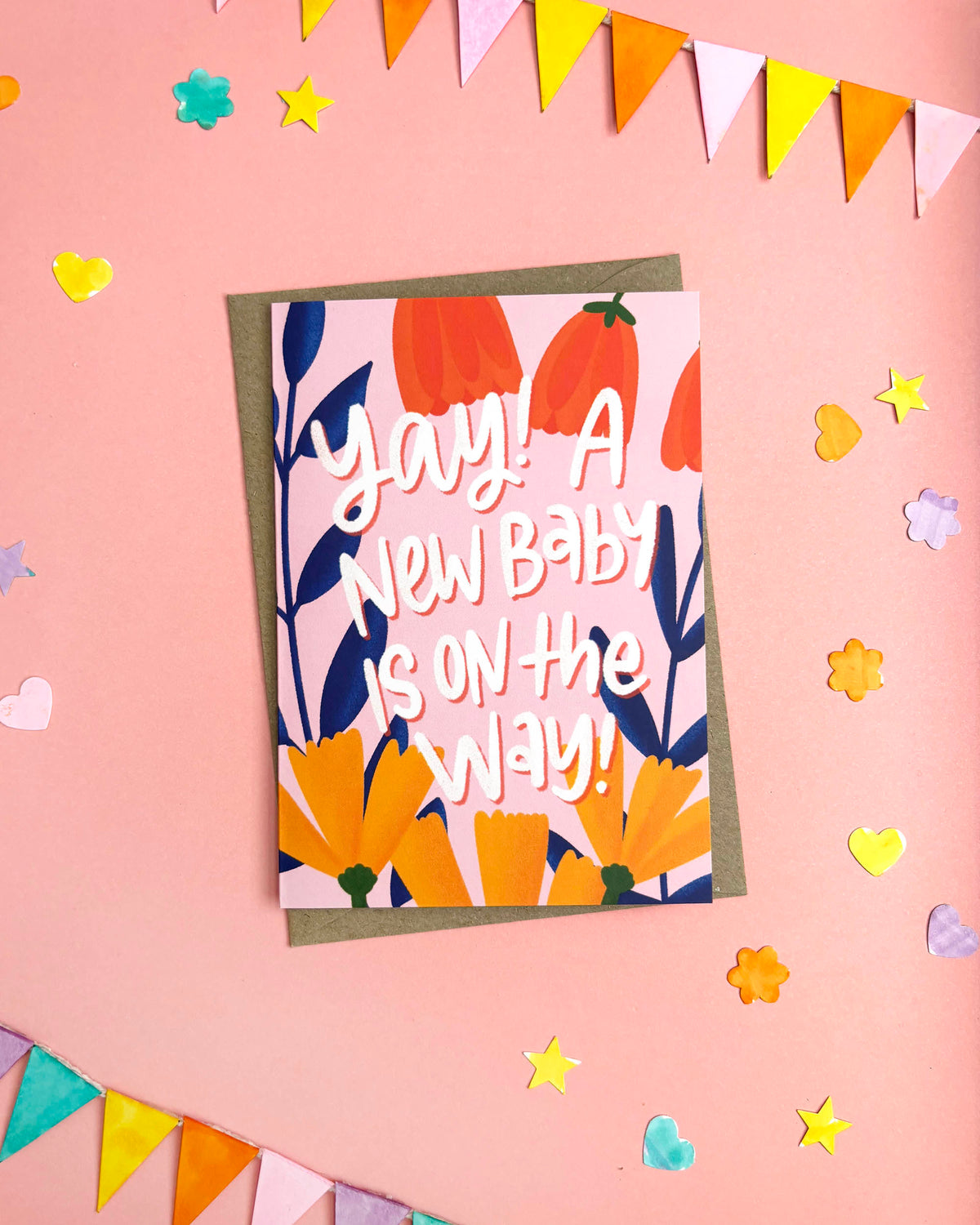 A colourful greeting card with a brown envelope, featuring the text 'yay! A NEW BABY IS ON THE WAY!' in white script over orange and navy floral illustrations. The card is displayed on a pink background decorated with bunting flags, hearts, and stars.