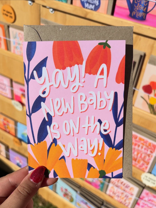 Card with floral design and text ' Yay! A new baby is on the way!' held in front of a display of greeting cards.