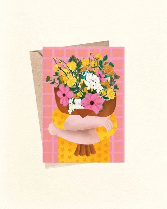 A square greeting card with a kraft envelope, featuring an illustration of a person hugging a large bouquet of yellow, pink, and white flowers against a pink plaid background.