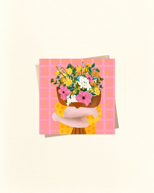 A square greeting card featuring an illustration of a person hugging a large bouquet of pink, yellow, and white flowers against a pink plaid background, shown with a matching envelope on a warm off-white surface.