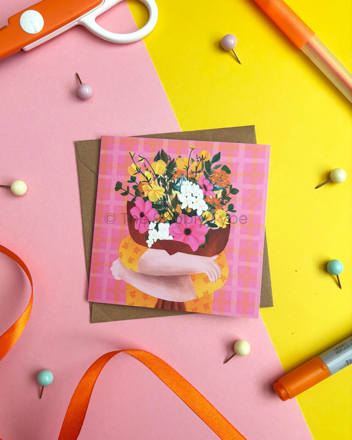 A square greeting card featuring a person hugging a colourful bouquet illustration on a pink plaid background, placed on top of a brown envelope. The card is photographed in a styled flat-lay arrangement on a split pink and yellow background with orange scissors, pins, ribbon, and markers around it.