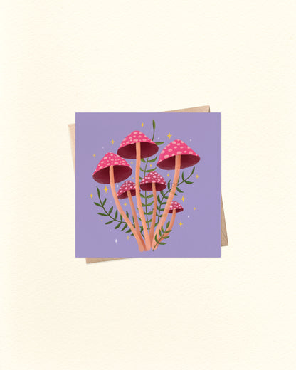 A square art print or card featuring a cluster of pink and red mushrooms with green stems on a lavender background, shown with a kraft envelope behind it on a warm off-white surface.