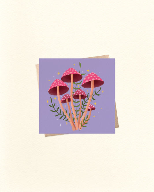 A square art print or card featuring a cluster of pink and red mushrooms with green stems on a lavender background, shown with a kraft envelope behind it on a warm off-white surface.
