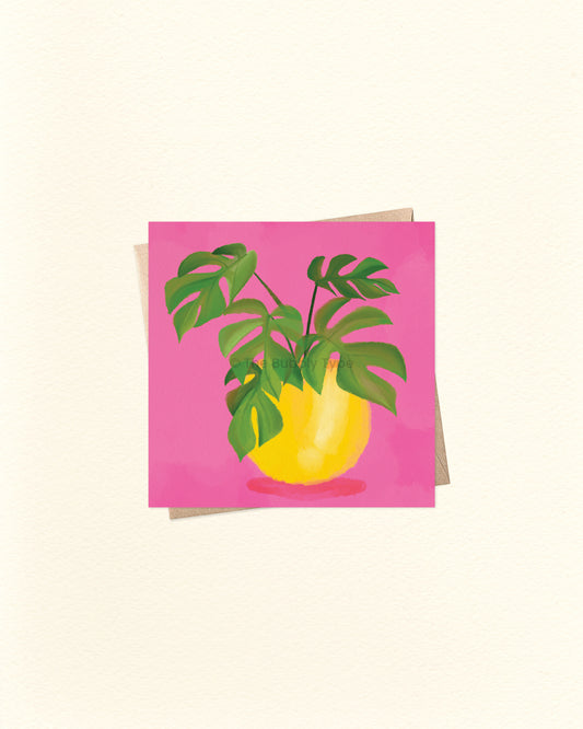 A square card featuring a stylised illustration of a yellow vase with green monstera leaves against a bright pink background, shown on a light cream textured surface with an envelope partially visible behind it.