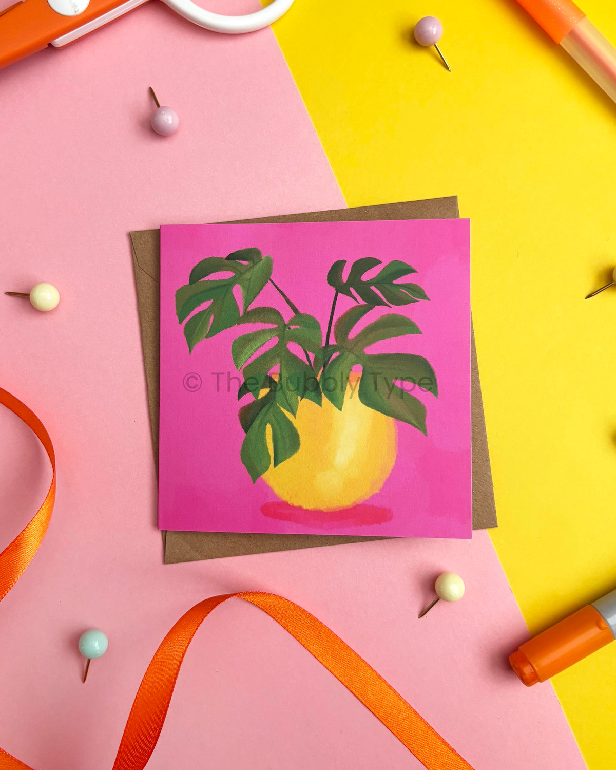 A square greeting card featuring a green monstera plant in a yellow pot against a bright pink background, styled on a brown envelope over a split pink and yellow surface with ribbon and pins.