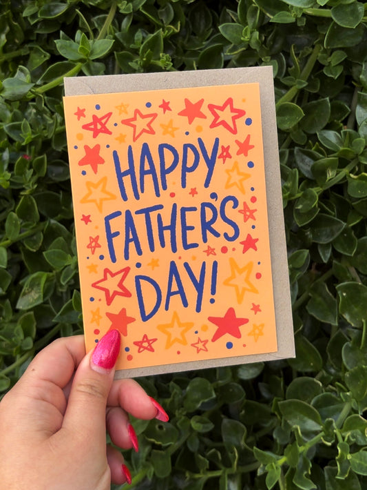 A hand with pink nail polish holds a square greeting card that says 'HAPPY FATHER'S DAY!' in blue lettering, surrounded by red and yellow stars on a peach-orange background. The card is placed in front of dense green foliage.