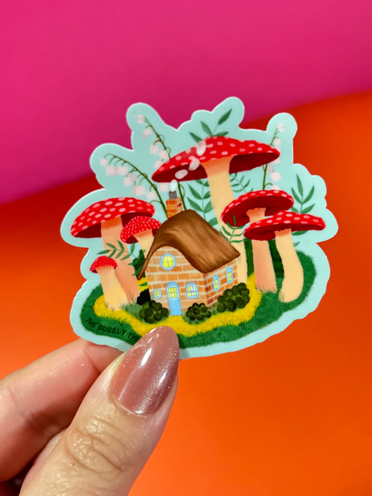 A hand with glittery nude nails holds a die-cut sticker featuring a whimsical mushroom forest with a small brown cottage. The sticker has a light blue border and is photographed against a vibrant pink and orange background.
