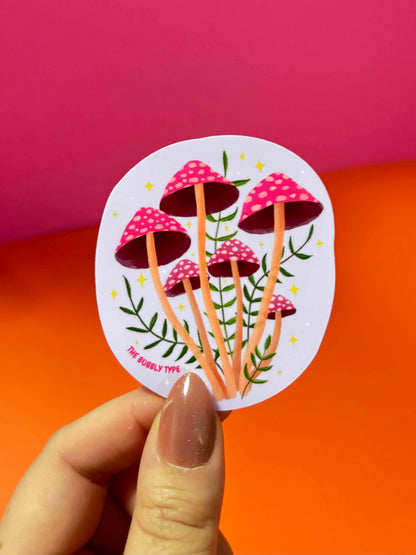 A hand with a nude manicure holds an imperfectly round sticker featuring a whimsical illustration of pink capped mushrooms with green leaves on a lavender background. The sticker is positioned against a vibrant, diagonally split pink and orange backdrop.