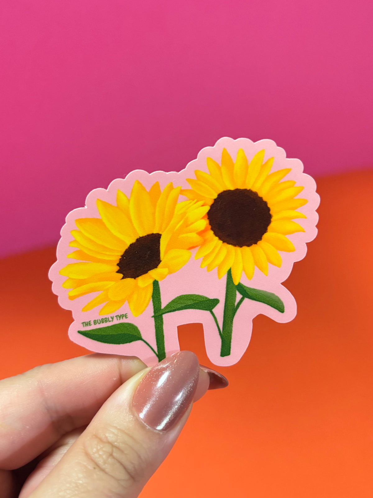 A hand with metallic pink nails holds a die-cut sticker featuring two yellow sunflowers with dark brown centres and green stems, set against a bright pink and orange background.