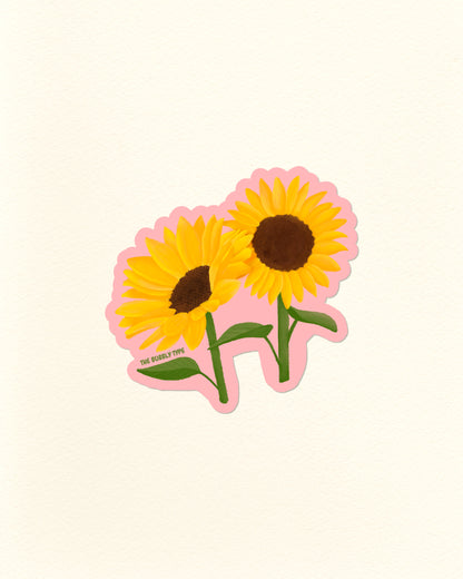 A die-cut sticker featuring two yellow sunflowers with brown centers, green stems, and a pink outline, shown on a plain off-white background. Small text near the lower left reads 'THE BUBBLY TYPE'.