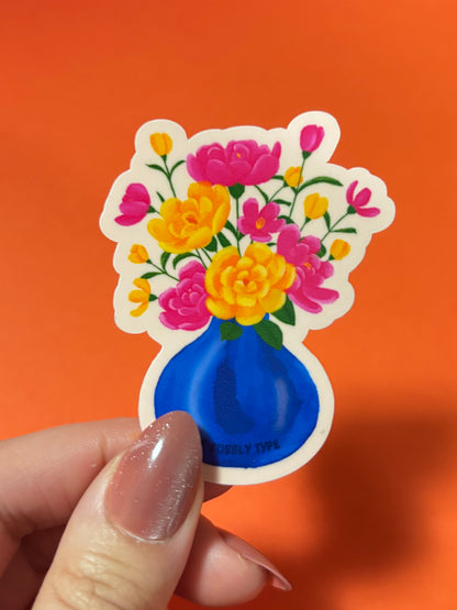 A hand with glittery nude nails holds a die-cut sticker featuring a blue vase filled with pink and yellow flowers, set against a solid orange background.