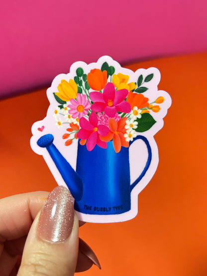 A hand with glittery nude nails holds a die-cut sticker featuring a blue watering can filled with colourful flowers, set against a bright pink and orange background.