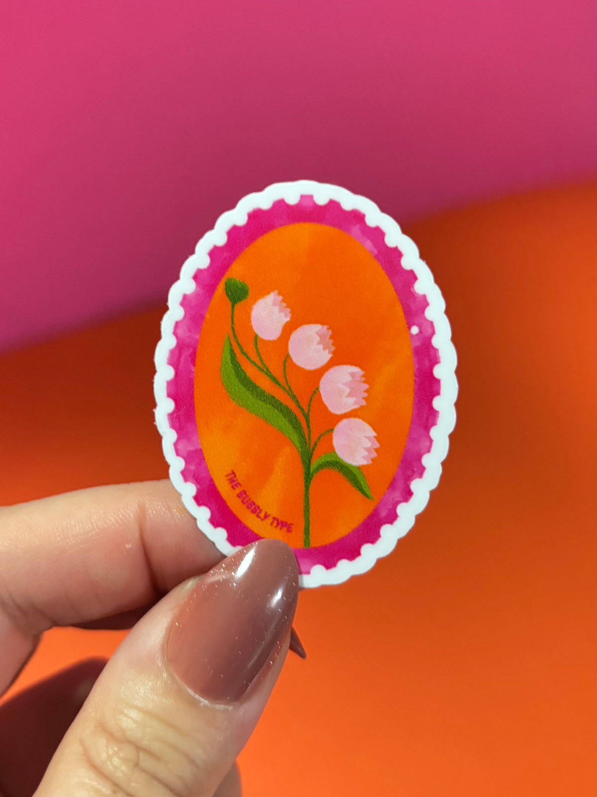 A hand with brown manicured nails holds a small oval sticker featuring a bright orange background, pink scalloped border, and a green plant with pink flowers. The background is a solid, vibrant orange.