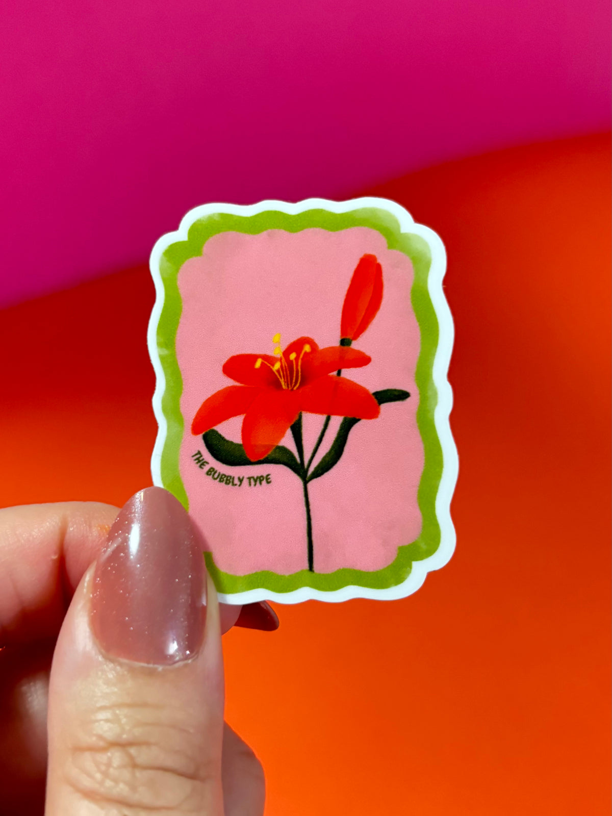 A hand with glittery nude nails holds a scalloped die-cut sticker featuring a red lily flower on a pink background with a green border. The sticker has the text 'THE BUBBLY TYPE' printed near the stem.