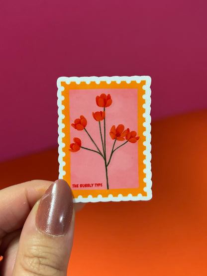 A hand with metallic mauve nail polish holds a small rectangular sticker featuring red flowers on a pink background with an orange border and white scalloped edge. The background is a solid gradient of magenta and orange.