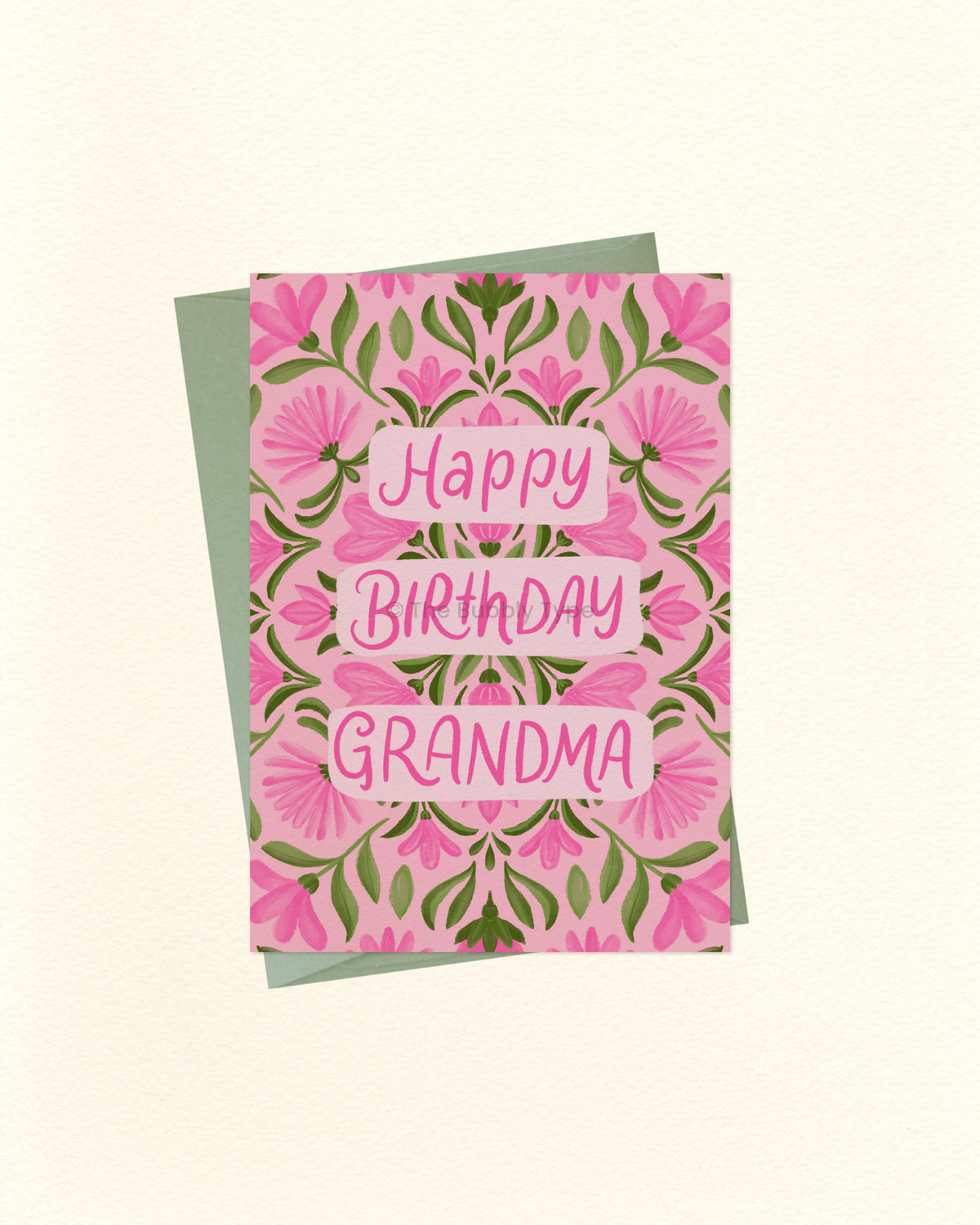 A floral 'Happy Birthday Grandma' greeting card with pink flowers and green leaves, layered over a sage green envelope on a warm off-white background.