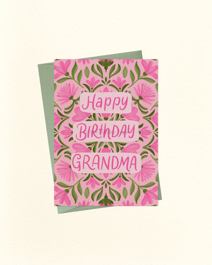 A floral 'Happy Birthday Grandma' greeting card with pink flowers and green leaves, layered over a sage green envelope on a warm off-white background.