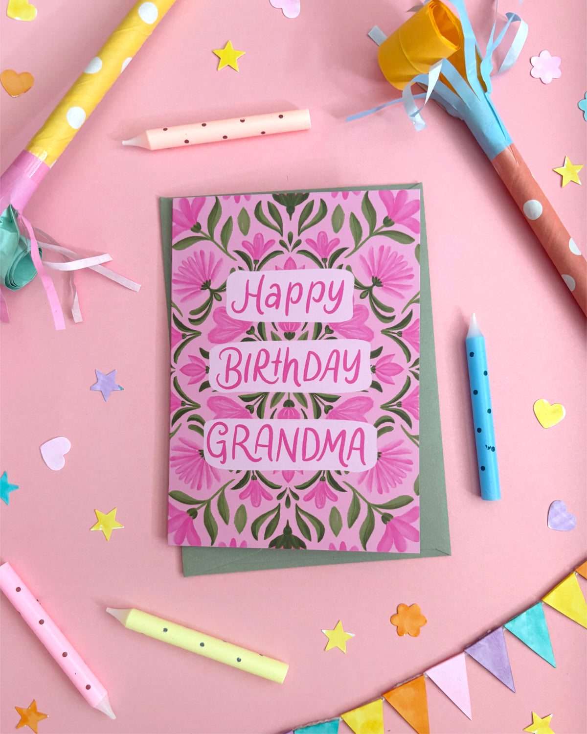An Australian-made birthday card for 'Grandma' featuring a pink and green floral damask pattern with 'Happy Birthday Grandma' written in pink letters. The card is styled in a flat-lay arrangement on a pink background with party props like candles, paper party hats, and star confetti.