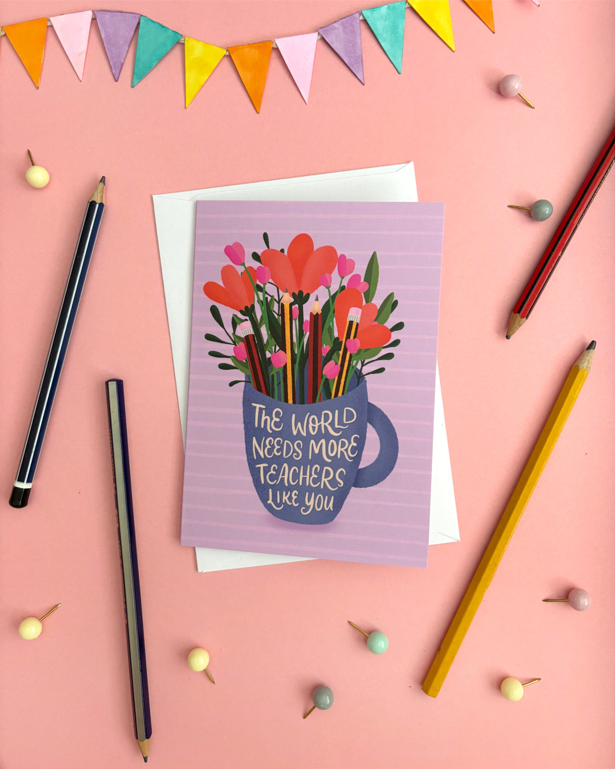 A greeting card with a purple striped background, featuring an illustration of orange and pink flowers in a blue mug with the text 'THE WORLD NEEDS MORE TEACHERS LIKE YOU'. The card is displayed on a pink background with greylead pencils, bunting, and small decorative pins.