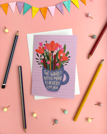 A greeting card with a purple striped background, featuring an illustration of orange and pink flowers in a blue mug with the text 'THE WORLD NEEDS MORE TEACHERS LIKE YOU'. The card is displayed on a pink background with greylead pencils, bunting, and small decorative pins.