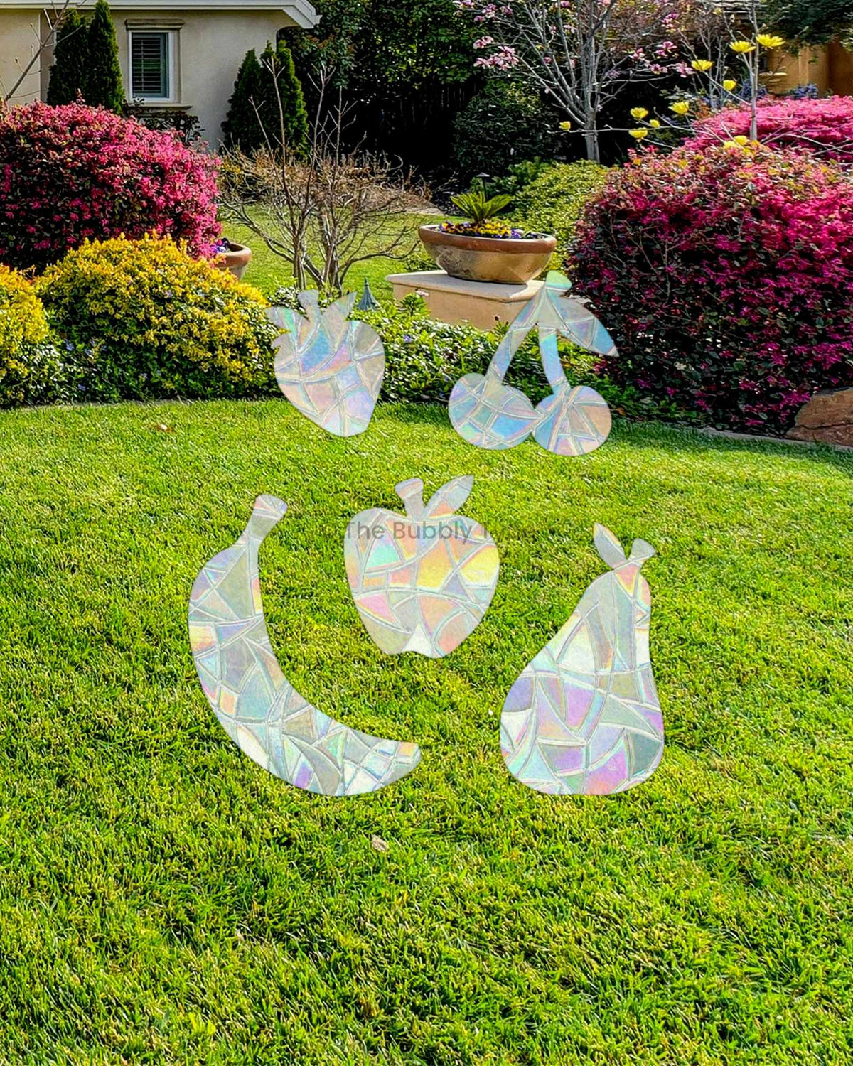 Iridescent holographic window stickers shaped like fruit, including a banana, pear, apple, and cherries, displayed on a window looking onto a green lawn in a landscaped garden setting.