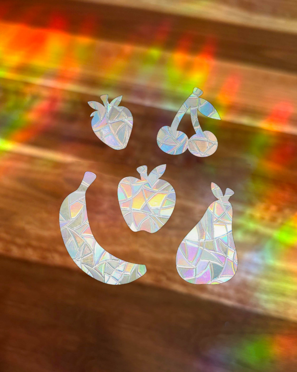 Five fruit-shaped window stickers or decals in strawberry, cherries, banana, apple, and pear shapes, decorated with a holographic, iridescent mosaic pattern, displayed on a wooden surface with colourful  rainbow light reflections.