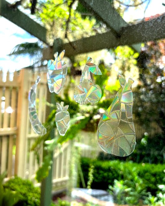 Iridescent fruit-themed sun catcher window sticker ornaments shaped like a pear, apple, cherry, strawberry and banana, displayed on a window in a sunny backyard garden setting.