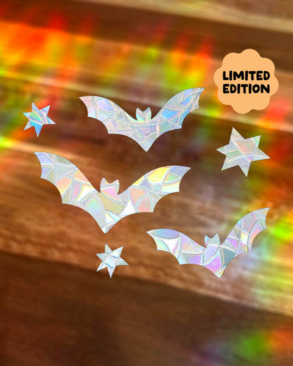 Three iridescent bat-shaped window stickers with star details are arranged on a wood surface with rainbow-prismatic lights shining. A badge in the top right reads 'LIMITED EDITION'.