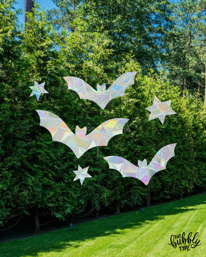 Five iridescent bat-shaped and star-shaped window decorations are displayed on a window looking onto a green backyard hedge and lawn. The holographic decorations reflect a rainbow of colours.
