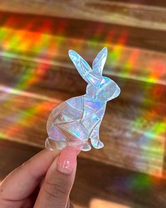 Iridescent rabbit-shaped sun catcher window sticker held by a hand with a colourful, rainbow lights, blurred background