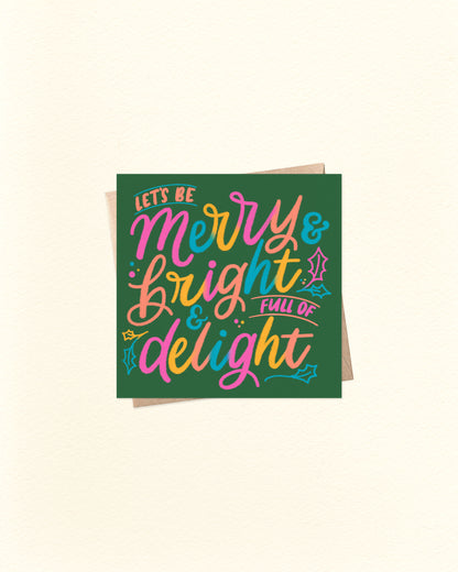 Christmas greeting card with colourful text reading "Let's Be Merry & Bright & Full of Delight" on a deep green background with brown kraft envelope.