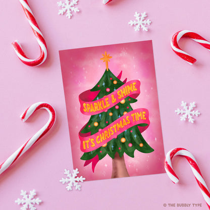 The same Christmas card laid flat on a pink background, surrounded by festive decorations including red and white candy canes and white snowflake paper cutouts