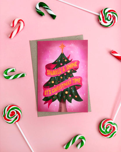 A Christmas greeting card featuring a stylized green Christmas tree with pink ribbons and the text 'SPARKLE & SHINE IT'S CHRISTMAS TIME' on a pink background, styled on a pink surface with candy canes and lollipops around it.