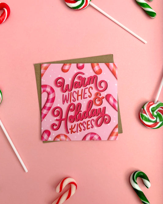 A square pink greeting card with the phrase 'Warm Wishes & Holiday Kisses' in hot pink script, layered over a brown envelope, arranged on a pink background with candy canes and lollipops around it.