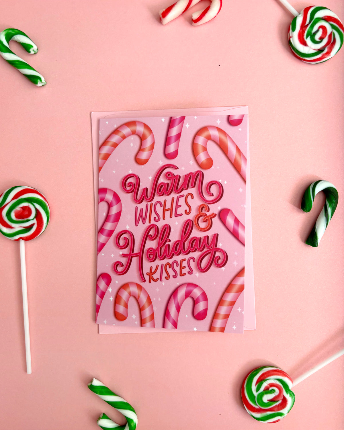 A pink greeting card with the words 'Warm Wishes & Holiday Kisses' in red script, surrounded by candy canes and lollipops on a pink background.