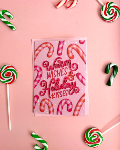 A pink greeting card with the words 'Warm Wishes & Holiday Kisses' in red script, surrounded by candy canes and lollipops on a pink background.