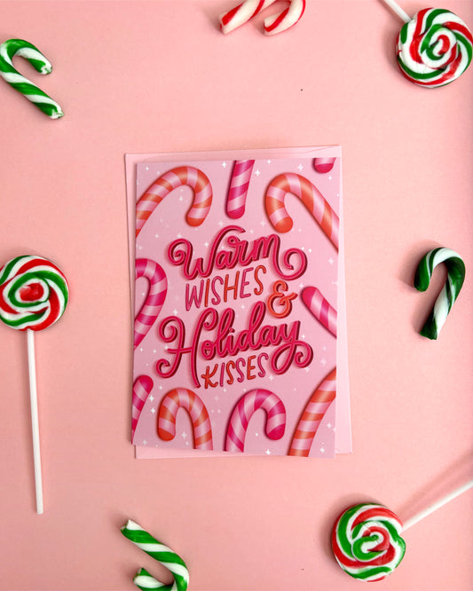 A pink greeting card with the words 'Warm Wishes & Holiday Kisses' in red script, surrounded by candy canes and lollipops on a pink background.