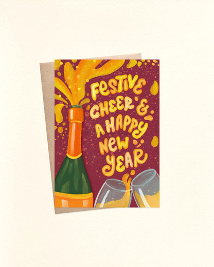 A6 Christmas card featuring an illustrated champagne bottle bursting with golden yellow champagne. The splash forms playful, hand-lettered words that read “Festive Cheer and Happy New Year.” Below are two champagne flutes clinking together, spilling bubbly into the air. The card is laid flat on top of a brown kraft envelope.