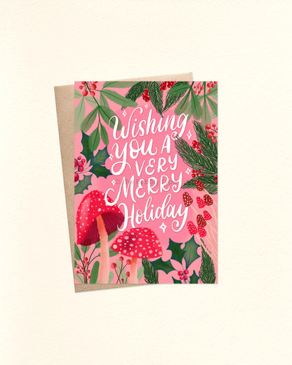 A6 Christmas card featuring white hand-lettered text that reads “Wishing You a Very Merry Holiday” on a pink background. Surrounding the text are hand-illustrated red-capped mushrooms of various shapes and sizes, green holly leaves, red berries, and festive foliage. The card is laid flat with a brown kraft envelope underneath.